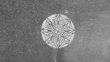 A detailed geometric pattern resembling a mandala is etched within a circle on a textured surface. The design features symmetrical lines and shapes converging into a central point, creating an intricate star-like appearance.