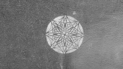 A detailed geometric pattern resembling a mandala is etched within a circle on a textured surface. The design features symmetrical lines and shapes converging into a central point, creating an intricate star-like appearance.