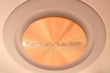 A close-up view of a round Harman Kardon speaker or audio component with a golden metallic finish, embedded in a circular frame. The texture and sheen of the material are visible, highlighting its premium build.