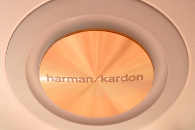 A close-up view of a round Harman Kardon speaker or audio component with a golden metallic finish, embedded in a circular frame. The texture and sheen of the material are visible, highlighting its premium build.