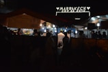 A nighttime street market scene with several people gathered under tents, illuminated by hanging bulbs. A sign above reads 'Marrakech COP 22 2016' suggesting an event or location. The atmosphere appears lively with food stalls and people wearing varied attire, some in chef uniforms.