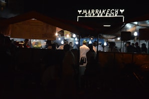 A nighttime street market scene with several people gathered under tents, illuminated by hanging bulbs. A sign above reads 'Marrakech COP 22 2016' suggesting an event or location. The atmosphere appears lively with food stalls and people wearing varied attire, some in chef uniforms.