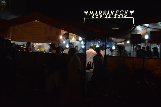 A nighttime street market scene with several people gathered under tents, illuminated by hanging bulbs. A sign above reads 'Marrakech COP 22 2016' suggesting an event or location. The atmosphere appears lively with food stalls and people wearing varied attire, some in chef uniforms.