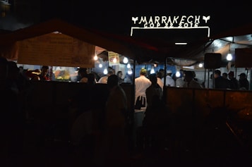A nighttime street market scene with several people gathered under tents, illuminated by hanging bulbs. A sign above reads 'Marrakech COP 22 2016' suggesting an event or location. The atmosphere appears lively with food stalls and people wearing varied attire, some in chef uniforms.