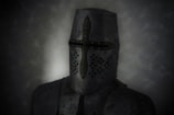 A medieval knight wearing a full face helmet stands against a dark, murky background. The helmet features a prominent cross-shaped design with small ventilation holes near the mouth area. The armor is made of metal and appears worn, with a slightly matte finish.