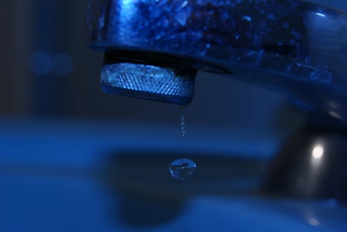 A close-up of a metal faucet with a single droplet of water falling from it, set against a blurred blue background.