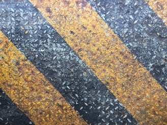 Diagonal black and yellow stripes on a rough, weathered surface, which appears to be asphalt or concrete, likely part of a road or pathway.