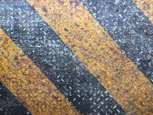 Diagonal black and yellow stripes on a rough, weathered surface, which appears to be asphalt or concrete, likely part of a road or pathway.