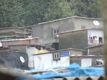 A collection of modest, stacked concrete and brick buildings with corrugated metal roofs. Many of the buildings feature faded or painted walls and some have satellite dishes. The structures appear worn and improvised. Surrounding the buildings are lush green trees.