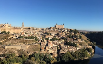 A panoramic view of a historic city situated on a hill, with a large river running alongside it. The skyline features prominent cathedrals and other ancient buildings with tiled rooftops. The landscape includes lush greenery and the architecture displays a blend of stone and brick structures.