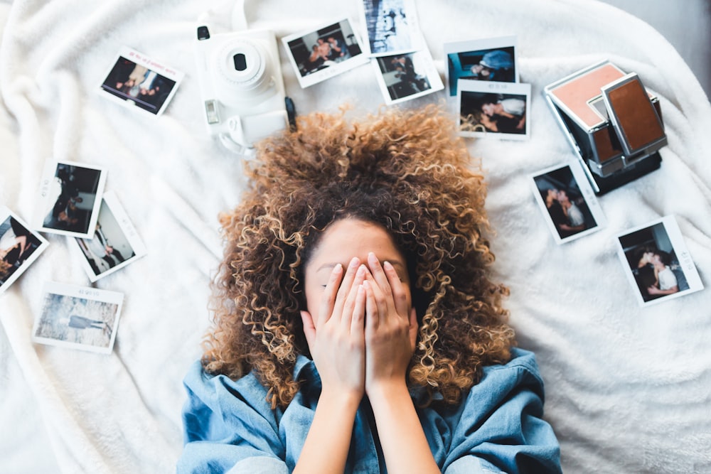 femme allongée sur un lit couvrant son visage entourée de photos et d’un appareil photo blanc