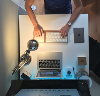 A neatly organized workspace features a person typing on a wireless keyboard. The desk holds a laptop, a tablet, a modern desk lamp, a notepad with a pen, a small box, and a cylindrical object. The setting has a minimalist and functional appeal, with wooden and metallic elements.