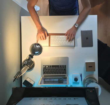 A neatly organized workspace features a person typing on a wireless keyboard. The desk holds a laptop, a tablet, a modern desk lamp, a notepad with a pen, a small box, and a cylindrical object. The setting has a minimalist and functional appeal, with wooden and metallic elements.