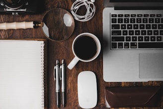 cup of coffee near MacBook Pro magnifying glass pens notebook and glasses