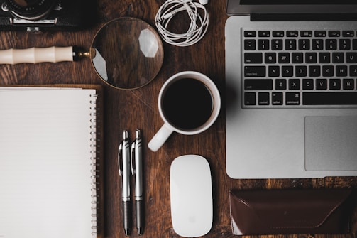 cup of coffee near MacBook Pro magnifying glass pens notebook and glasses