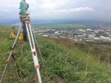 A surveyor's tripod with a total station is set up on a grassy hill overlooking a sprawling rural area with fields and industrial buildings. The sky is overcast, slightly obscuring the distant horizon, and there is a utility bag on the ground nearby.