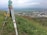 A surveyor's tripod with a total station is set up on a grassy hill overlooking a sprawling rural area with fields and industrial buildings. The sky is overcast, slightly obscuring the distant horizon, and there is a utility bag on the ground nearby.