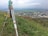 A surveyor's tripod with a total station is set up on a grassy hill overlooking a sprawling rural area with fields and industrial buildings. The sky is overcast, slightly obscuring the distant horizon, and there is a utility bag on the ground nearby.
