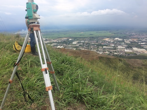 A surveyor's tripod with a total station is set up on a grassy hill overlooking a sprawling rural area with fields and industrial buildings. The sky is overcast, slightly obscuring the distant horizon, and there is a utility bag on the ground nearby.