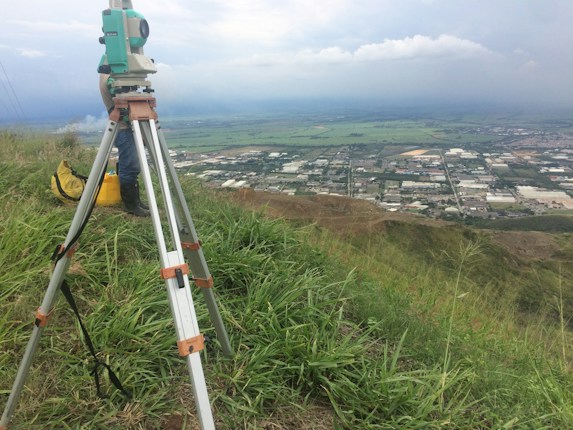 A surveyor's tripod with a total station is set up on a grassy hill overlooking a sprawling rural area with fields and industrial buildings. The sky is overcast, slightly obscuring the distant horizon, and there is a utility bag on the ground nearby.