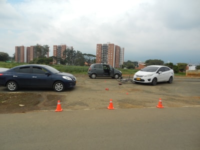A roadside scene with two cars, one blue and one white, parked near a bicycle that appears to be involved in a minor accident. Traffic cones are placed around the area, and residential buildings are visible in the background under a cloudy sky.
