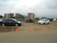 A roadside scene with two cars, one blue and one white, parked near a bicycle that appears to be involved in a minor accident. Traffic cones are placed around the area, and residential buildings are visible in the background under a cloudy sky.