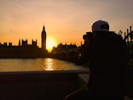 A photographer capturing the sunset over the iconic Big Ben and Houses of Parliament in London. The scene is set by a tranquil river with the sun casting an orange glow across the sky and water. A lone bird flies near the towering silhouette of Big Ben.