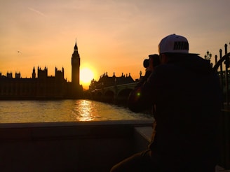 A photographer capturing the sunset over the iconic Big Ben and Houses of Parliament in London. The scene is set by a tranquil river with the sun casting an orange glow across the sky and water. A lone bird flies near the towering silhouette of Big Ben.