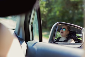 Reflection in a car's side mirror of a person wearing sunglasses, smiling, and possibly singing. The scene is bright, with lush green foliage in the background.