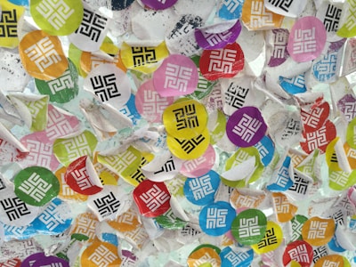 A chaotic arrangement of colorful, circular stickers with geometric black and white patterns, some partially peeled off a textured surface. The circles come in various colors including blue, red, yellow, green, and purple.