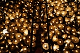 A multitude of glowing circular lamps are clustered together, creating a warm, golden ambiance. The lamps are arranged in a dense pattern with a lattice-like structure running vertically through the center. The light emitted is soft and diffused, casting a serene glow across the scene.