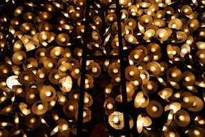 A multitude of glowing circular lamps are clustered together, creating a warm, golden ambiance. The lamps are arranged in a dense pattern with a lattice-like structure running vertically through the center. The light emitted is soft and diffused, casting a serene glow across the scene.