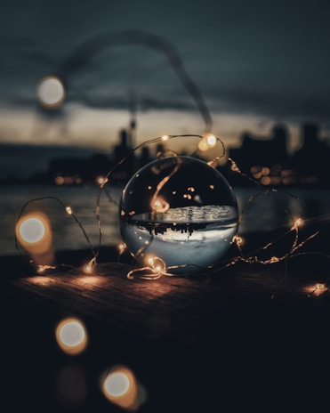 A crystal ball placed on a wooden surface reflects a city skyline at dusk. Surrounding the ball, soft, warm fairy lights create a bokeh effect against the dark background. The sky appears moody and overcast, enhancing the magical ambiance.