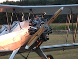 A vintage biplane with a wooden propeller is parked on a grassy field. The aircraft features a polished metal exterior with visible engine components and German text on the fuselage. The backdrop includes a cornfield and a dense forest area, with a few people visible in the distance.