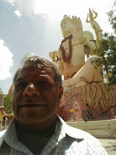 A large statue of a deity in a sitting posture, adorned with traditional ornaments and holding a trident, dominates the scene. A man is taking a selfie in the foreground, capturing the statue and other people walking around the area. The sky is partly cloudy and the surroundings include trees and a few buildings.