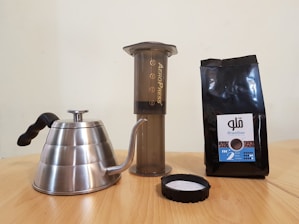 A stainless steel gooseneck kettle, an Aeropress coffee maker, and a bag of Brazilian coffee beans are arranged on a wooden surface. The kettle features a black handle, and the coffee bag is dark with a label.