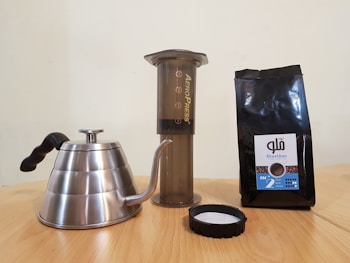 A stainless steel gooseneck kettle, an Aeropress coffee maker, and a bag of Brazilian coffee beans are arranged on a wooden surface. The kettle features a black handle, and the coffee bag is dark with a label.