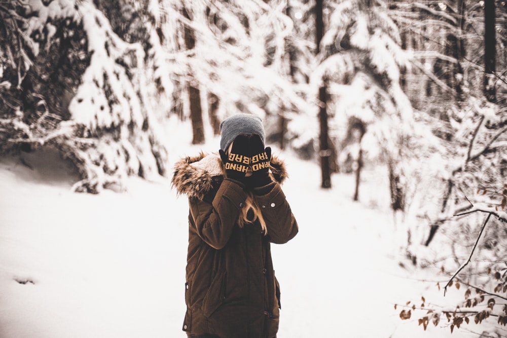 person standing on snow field surrounded by trees covering his face with his hands