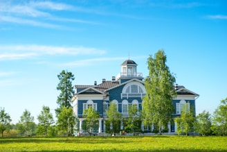A large, two-story blue building with white trim is set in a lush, green landscape. Surrounding the building are several tall trees and a well-maintained lawn. The sky is clear and blue, adding to the serene atmosphere.