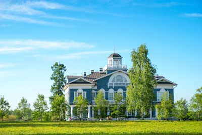 A large, two-story blue building with white trim is set in a lush, green landscape. Surrounding the building are several tall trees and a well-maintained lawn. The sky is clear and blue, adding to the serene atmosphere.