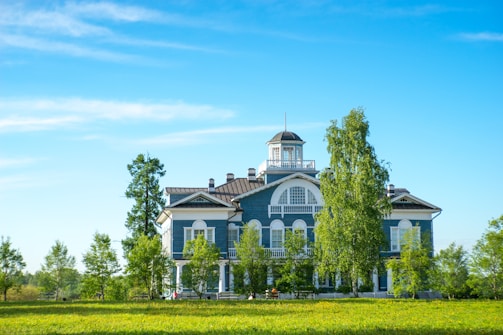 A large, two-story blue building with white trim is set in a lush, green landscape. Surrounding the building are several tall trees and a well-maintained lawn. The sky is clear and blue, adding to the serene atmosphere.