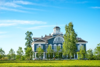 A large, two-story blue building with white trim is set in a lush, green landscape. Surrounding the building are several tall trees and a well-maintained lawn. The sky is clear and blue, adding to the serene atmosphere.