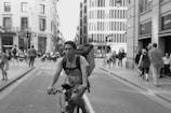 A cyclist riding in an urban street surrounded by pedestrians. The street is lined with tall buildings, and there is a bustling atmosphere with people walking and scooters parked nearby. The cyclist, who appears focused, is wearing a backpack and navigating through the traffic.
