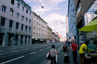 A street scene with multiple people walking along a sidewalk beside several buildings, including one with a sign reading 'Raiffeisenbank.' The road is lined with overhead wires and appears to be relatively empty of vehicles. Shops and signage are visible on both sides of the street, and the sky is partly cloudy.
