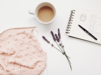 A cozy scene with a pink knitted fabric on knitting needles, a white mug filled with coffee or tea, a sketchbook with handwritten notes and a knitting pattern diagram, a black pen, and a small bunch of lavender flowers laid on a white background.