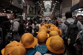 A bustling indoor marketplace is filled with people, including a group of children wearing bright yellow hats. The market has various stalls along the sides and banners hanging above. The combination of the crowd and vibrant colors creates a lively atmosphere.