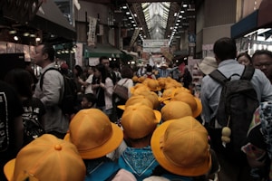 A bustling indoor marketplace is filled with people, including a group of children wearing bright yellow hats. The market has various stalls along the sides and banners hanging above. The combination of the crowd and vibrant colors creates a lively atmosphere.