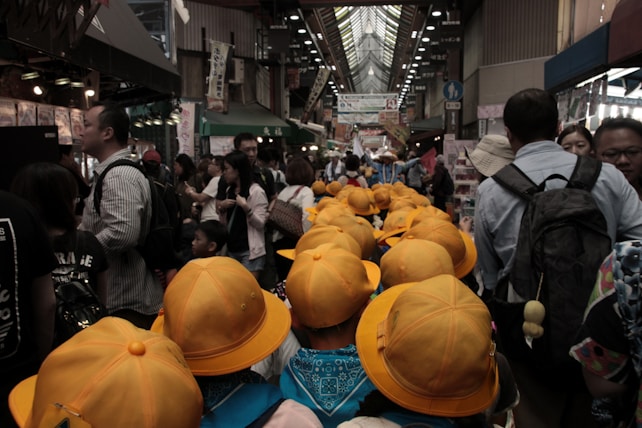 A bustling indoor marketplace is filled with people, including a group of children wearing bright yellow hats. The market has various stalls along the sides and banners hanging above. The combination of the crowd and vibrant colors creates a lively atmosphere.