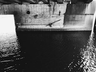 A concrete bridge structure cast in shadow with rippling water below. The image is in black and white, emphasizing the textures and contrasts of the rough concrete and the smooth water surface. Light streams in from the left, creating a stark contrast and highlighting parts of the structure.