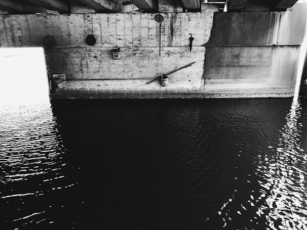 A concrete bridge structure cast in shadow with rippling water below. The image is in black and white, emphasizing the textures and contrasts of the rough concrete and the smooth water surface. Light streams in from the left, creating a stark contrast and highlighting parts of the structure.